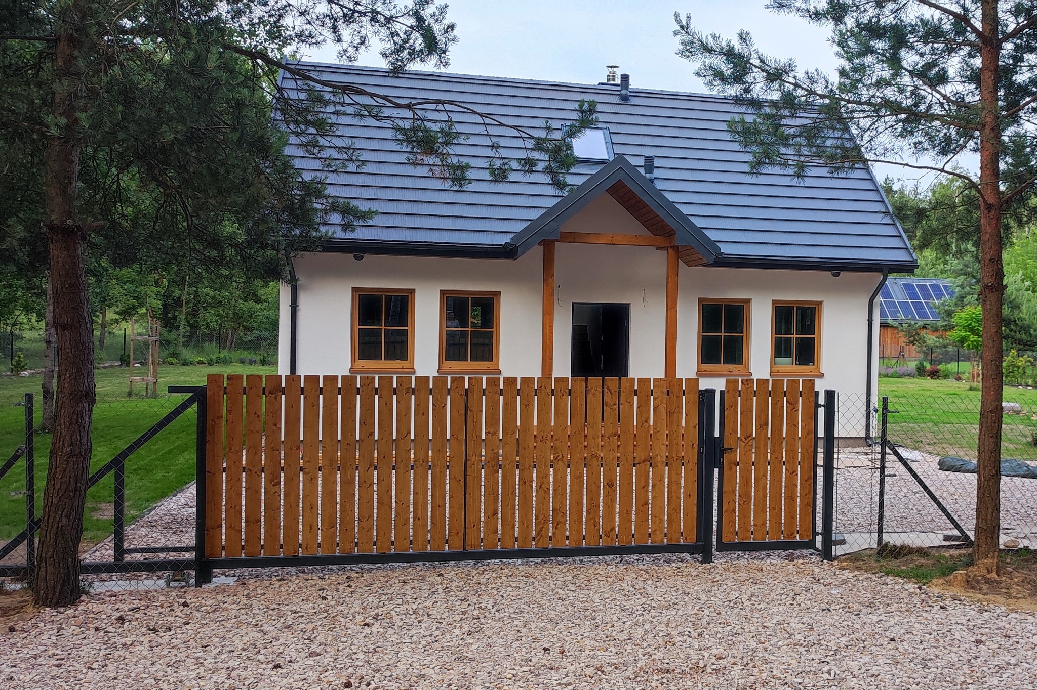 Nowoczesny dom szkieletowy z drewnianym ogrodzeniem panelowym i szarą dachówką, widok z zewnątrz na posesję wyłożoną żwirem.