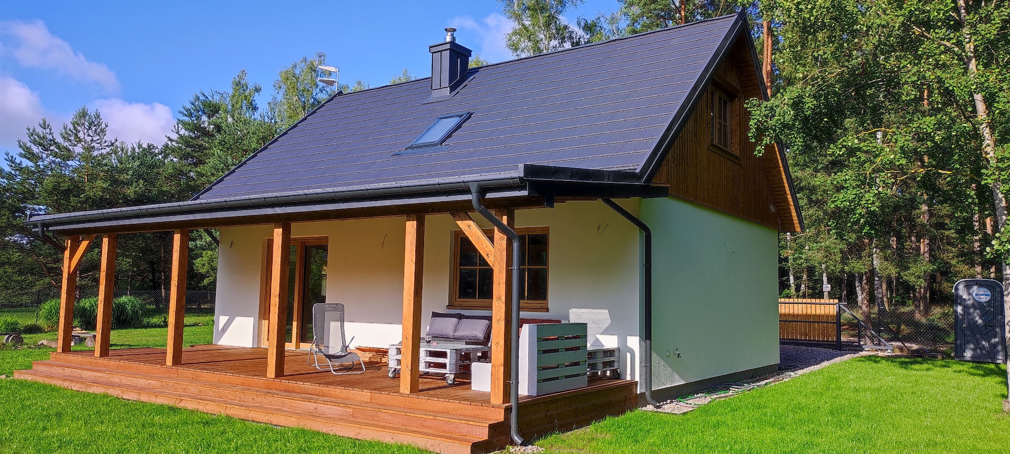 Gotowy, parterowy dom z szarą dachówką i drewnianym tarasem, otoczony zielenią, widoczny przenośny wychodek Toi Toi.