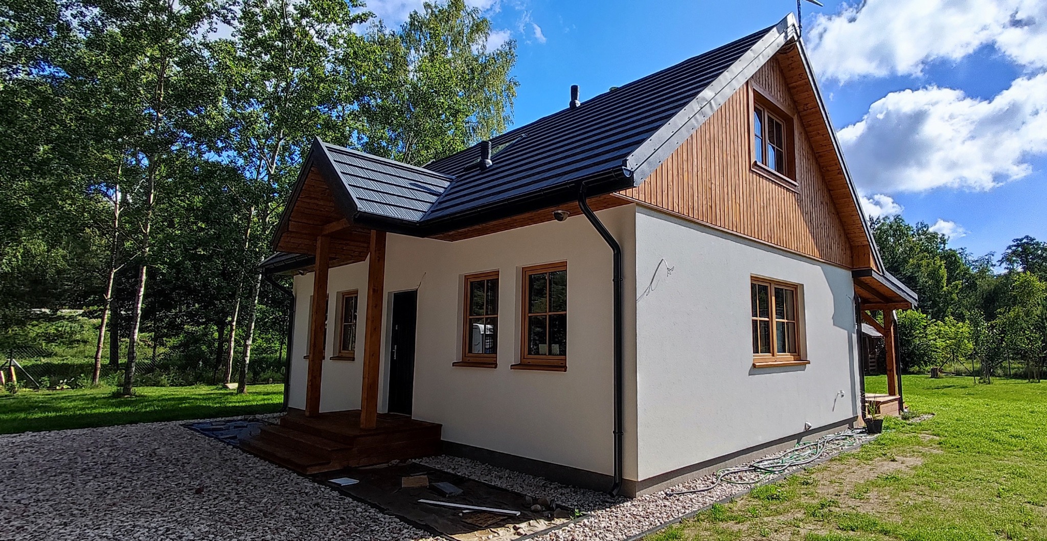 Gotowy dom z drewnianą elewacją na poddaszu i ciemnym dachem, otoczony zielenią i jasnym żwirem.
