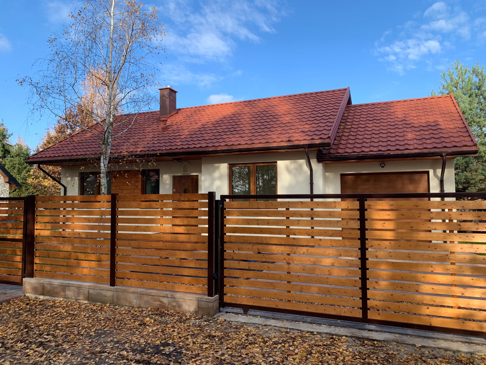 Gotowy dom jednorodzinny z czerwoną dachówką, ogrodzony drewnianym płotem z poziomych desek, widoczny jesienny krajobraz z opadłymi liśćmi.