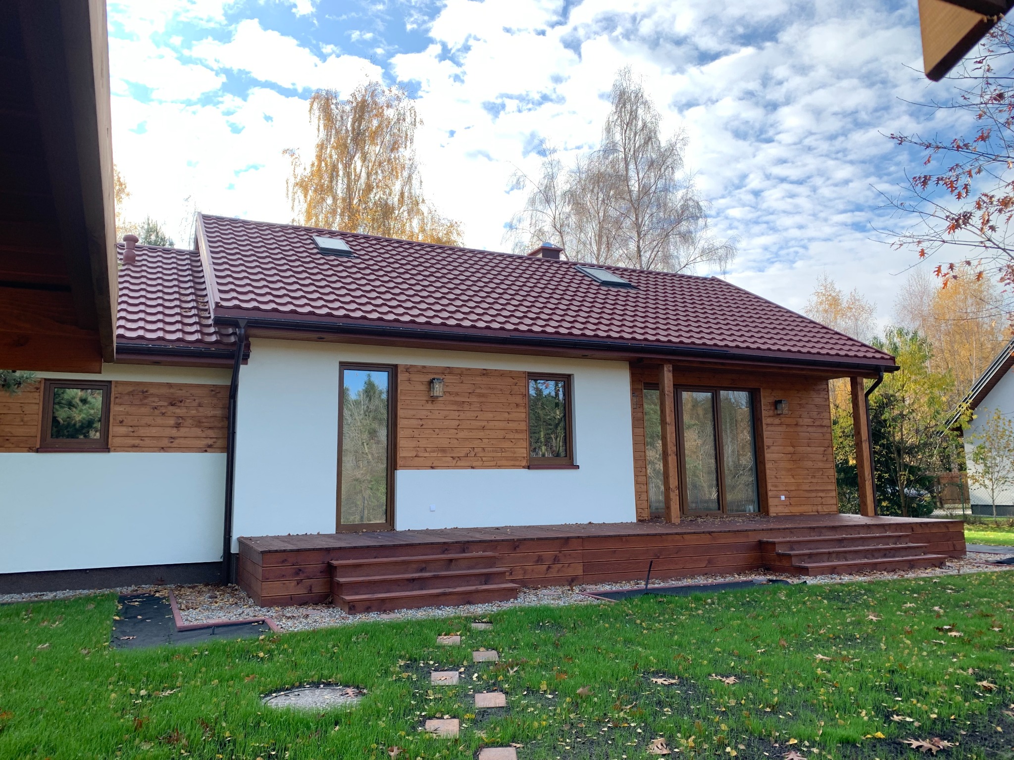 Nowoczesny dom z drewnianą elewacją i czerwonym dachem, otoczony zielonym trawnikiem z kamienną ścieżką, widok z poziomu gruntu w słoneczny dzień.