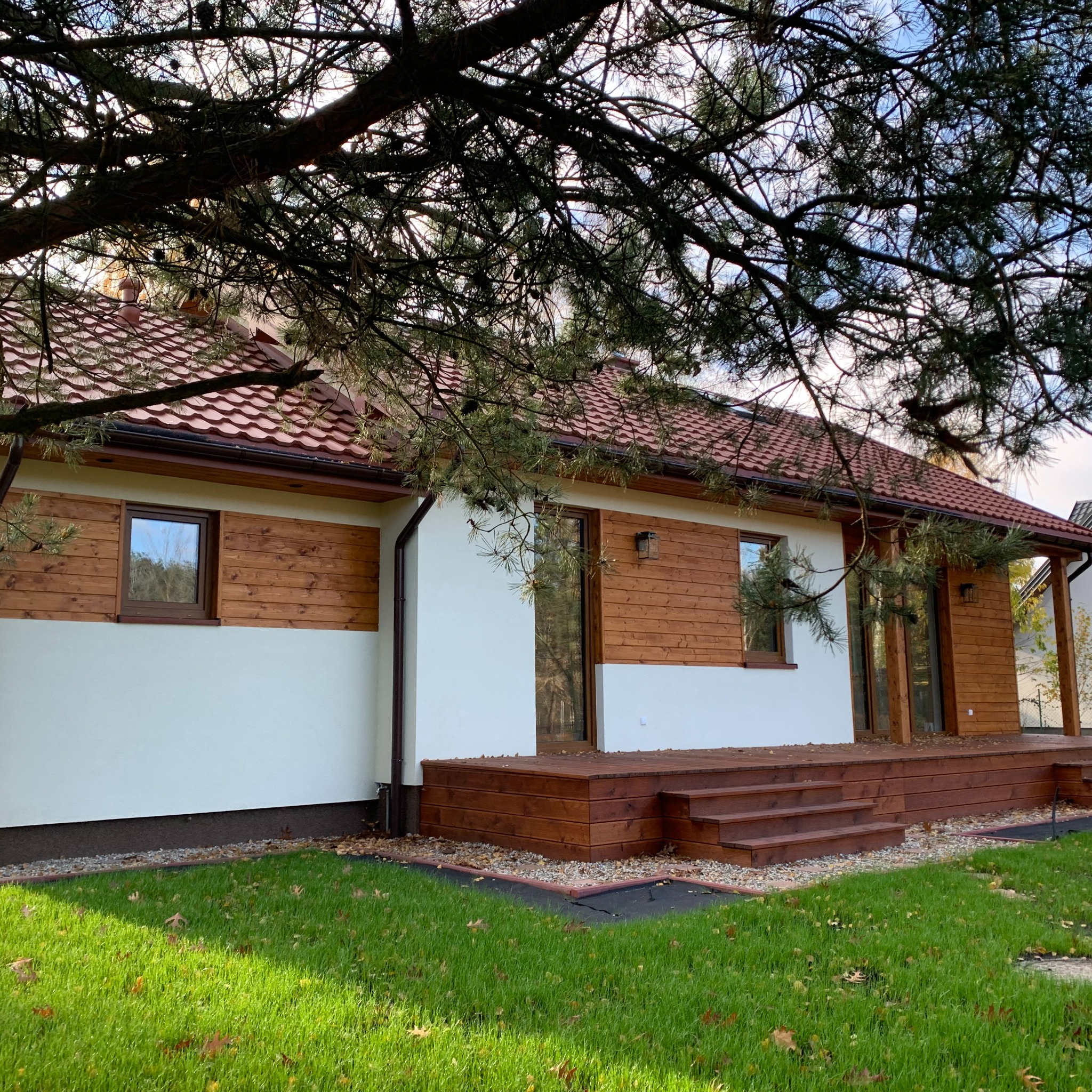 Parterowy dom z drewnianą elewacją i czerwonym dachem, widoczny z perspektywy trawnika, częściowo zasłonięty gałęziami drzewa.