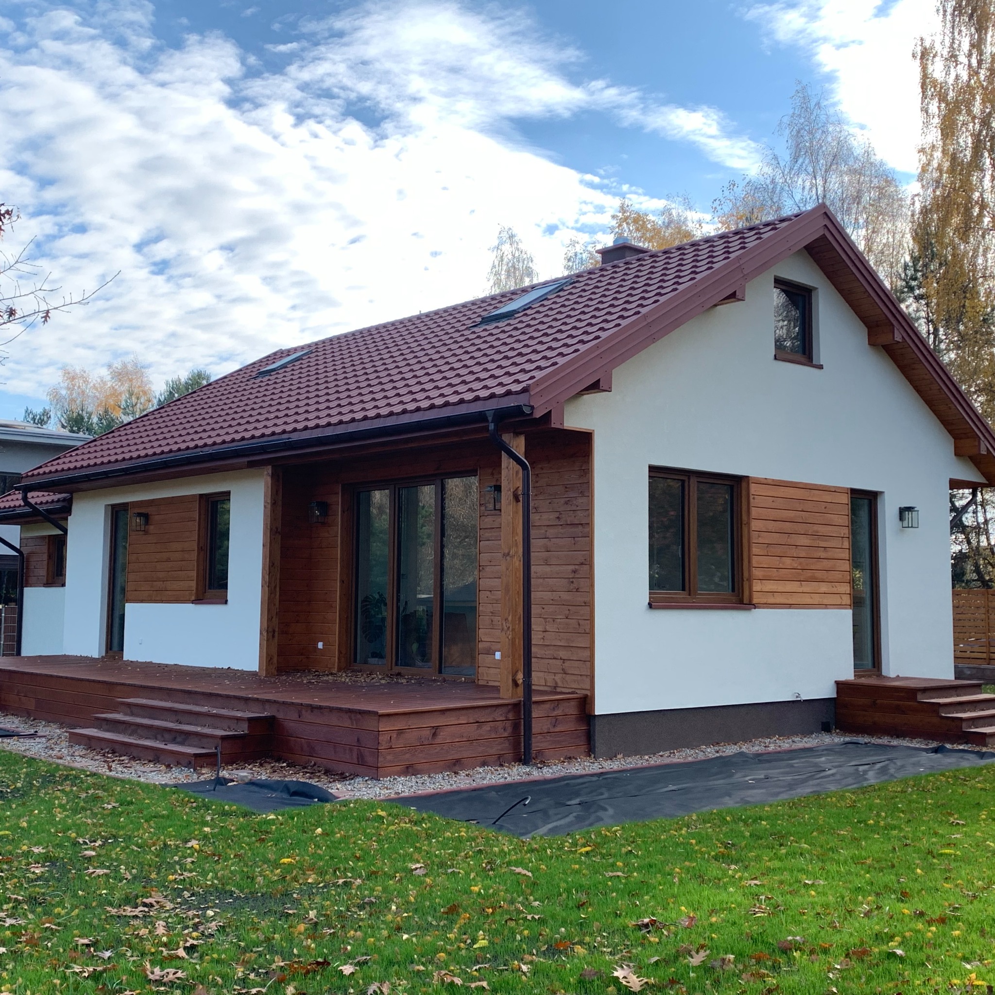 Gotowy dom szkieletowy z drewnianym tarasem i brązową dachówką, otoczony zielonym trawnikiem z opadłymi liśćmi.
