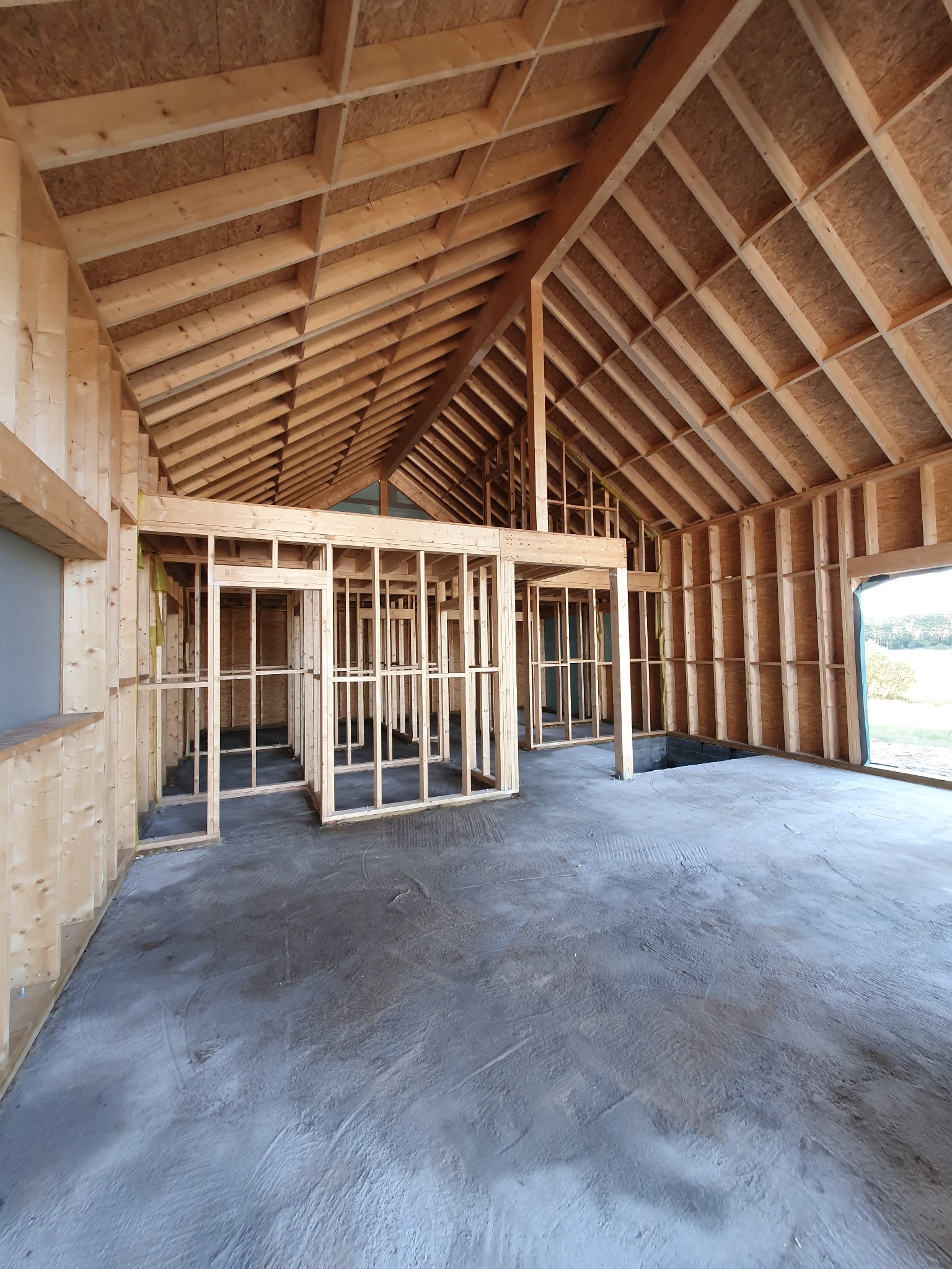 Wnętrze w trakcie budowy z drewnianą konstrukcją szkieletową ścian i dachu, surowy beton na podłodze, widok na zewnątrz przez otwór okienny.