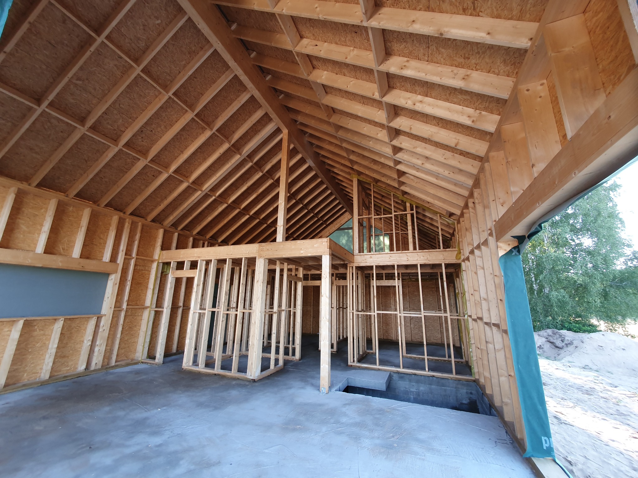 Wnętrze domu w trakcie budowy, widoczne drewniane szkielety ścian i dachu, surowy beton na podłodze, przygotowane otwory w podłodze, widok na zewnątrz przez niezabudowane ściany.