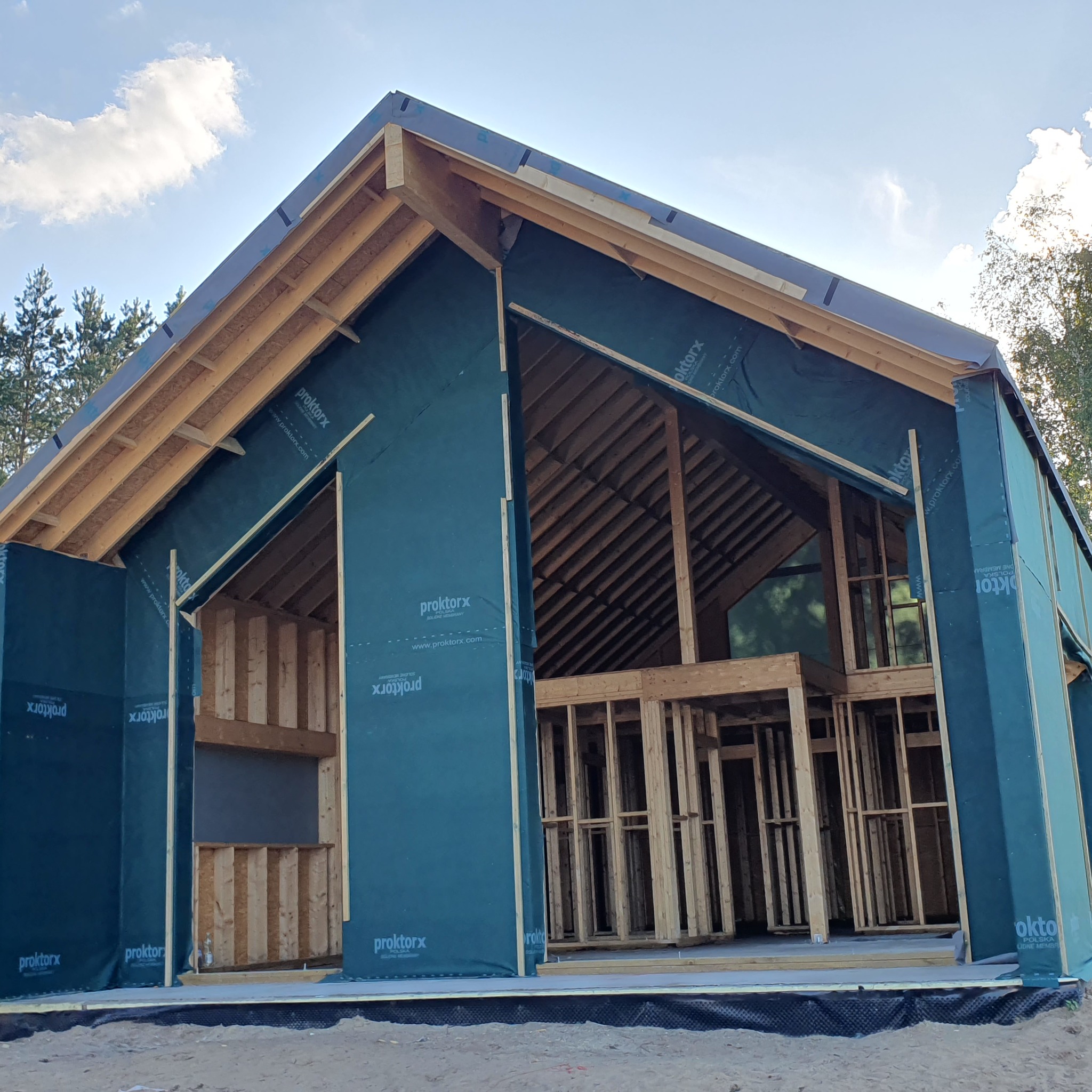 Konstrukcja domu w trakcie budowy, widoczne drewniane ramy ścian i dachu pokryte zieloną membraną ochronną, z widocznym niebem i drzewami w tle.