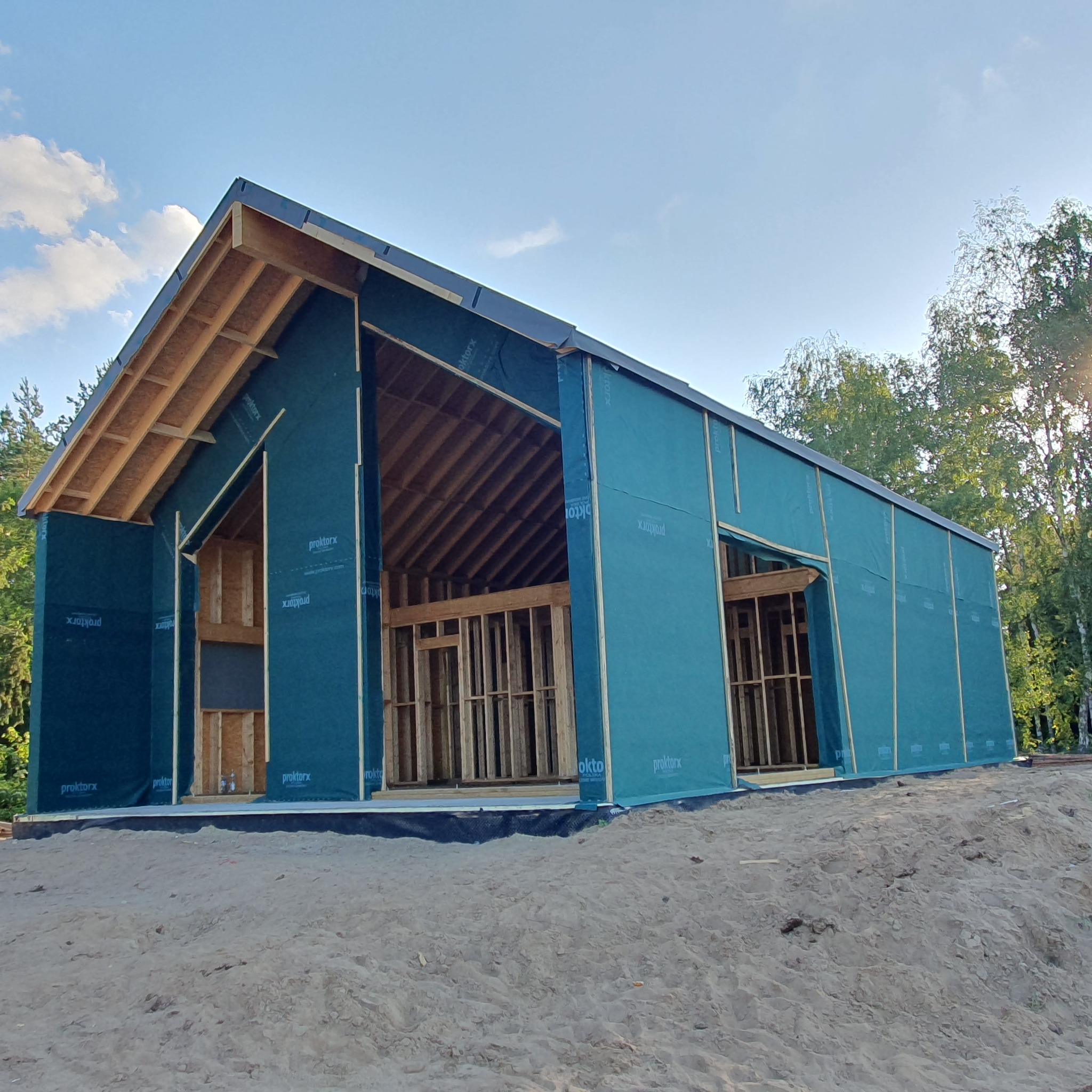 Surowa konstrukcja domu z drewnianą ramą, pokryta niebieską membraną ochronną z widocznym napisem 'proktorx', na piaszczystym terenie, z lasem w tle i błękitnym niebem.