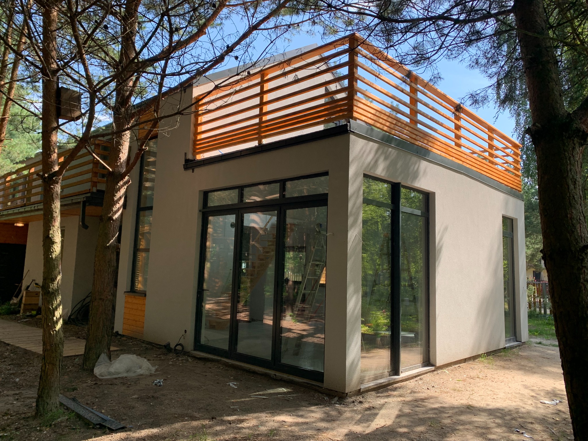 Nowoczesny dom szkieletowy z płaskim dachem i drewnianą balustradą na dachu, duże okna, widok z zewnątrz w otoczeniu drzew.