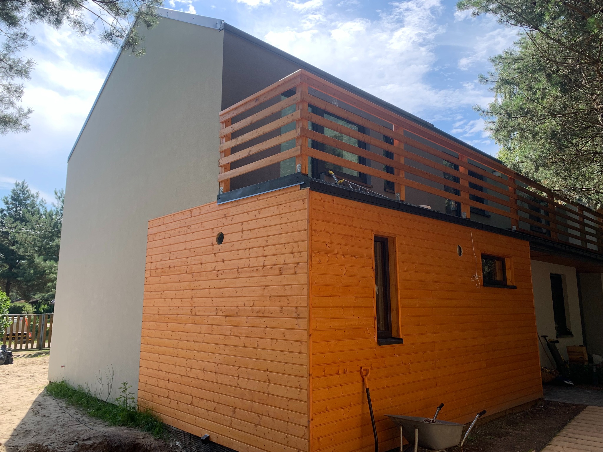 Nowoczesny dom z drewnianą elewacją i balkonem z drewnianą balustradą, widoczny wózek budowlany i łopata, częściowo otynkowana ściana, błękitne niebo w tle.