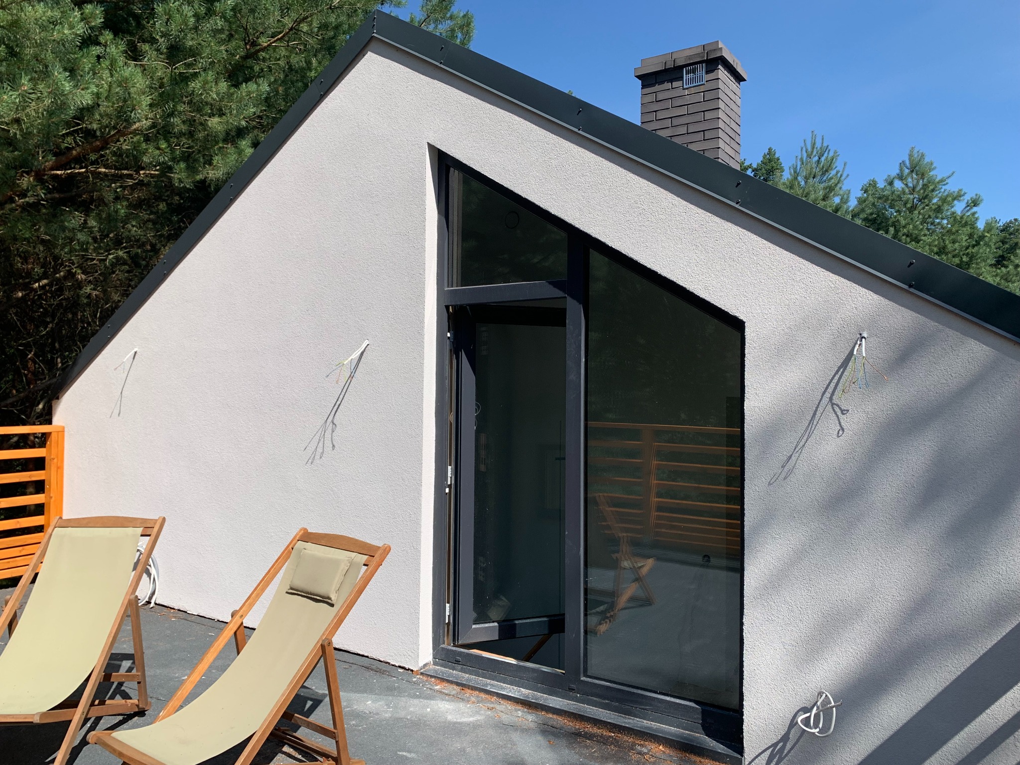 Nowoczesny budynek z jasną elewacją i skośnym dachem, z dużym trójkątnym oknem i otwartymi drzwiami na taras, na którym stoją dwa leżaki.