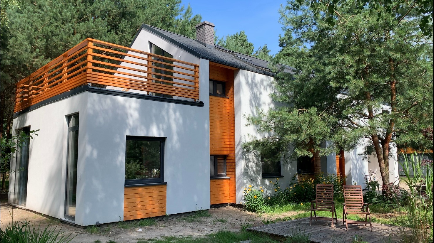 Nowoczesny dom z białym tynkiem i drewnianą elewacją, balkon z drewnianą balustradą, otoczony zielenią i drzewami, widok z zewnątrz w słoneczny dzień.