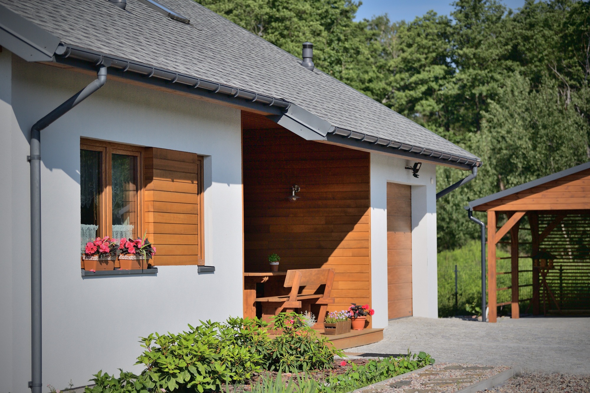Nowoczesny dom z białymi ścianami i drewnianymi elementami, okna z kwiatami, zadaszone drewniane patio z meblami ogrodowymi, widok na ogród i drewutnię.