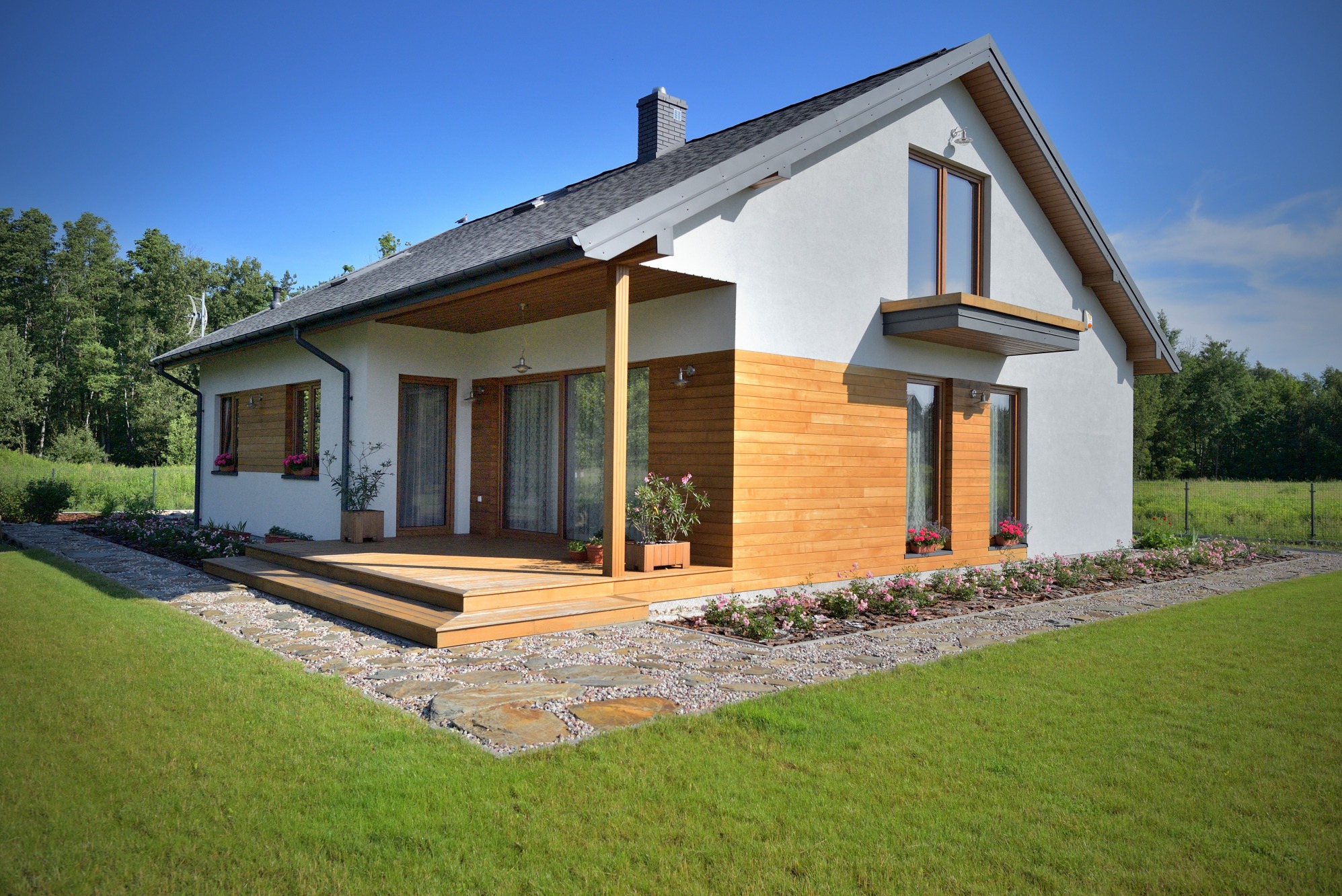 Nowoczesny dom z elementami drewnianymi, tarasem i zadbanym ogrodem w słoneczny dzień. Widoczna drewniana elewacja, szara dachówka i starannie ułożony kamienny chodnik.