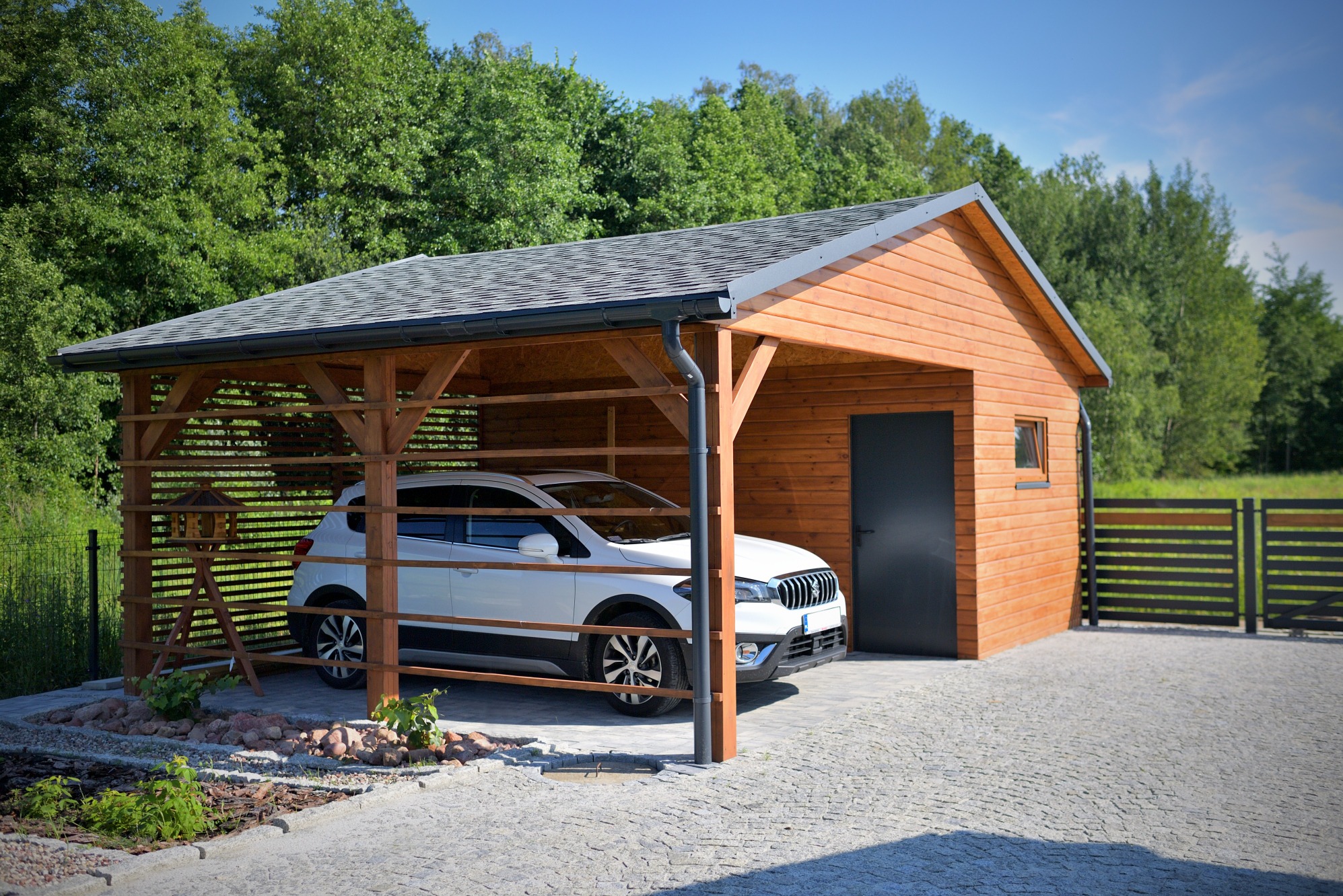 Drewniany garaż z zadaszeniem i ścianą boczną, częściowo otwarty, z białym samochodem w środku, na tle zieleni.