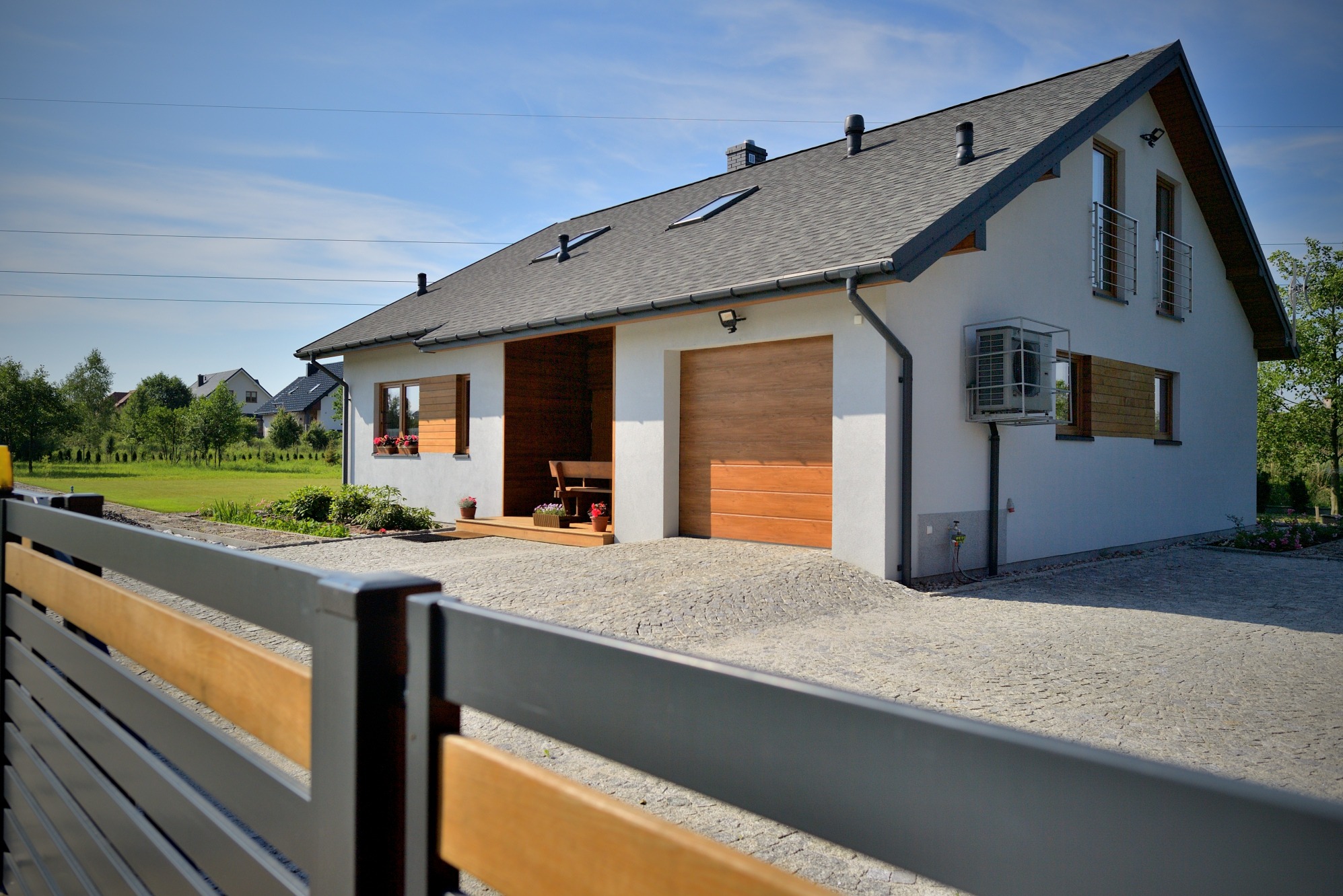 Nowoczesny dom jednorodzinny z garażem, wykończony w jasnych kolorach, z drewnianymi akcentami i zadbanym otoczeniem, widziany zza ogrodzenia.