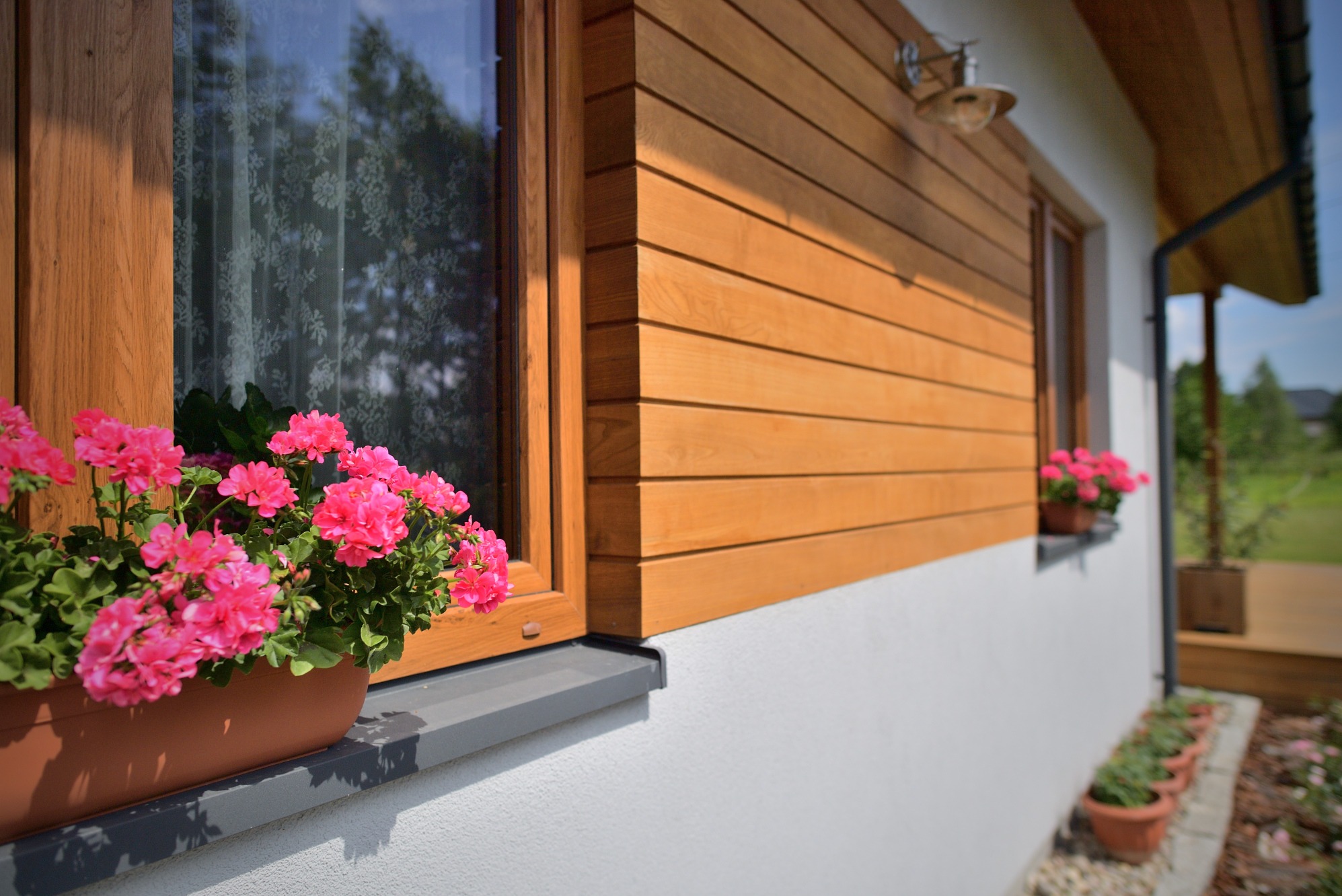 Fragment domu z drewnianą elewacją, oknem z drewnianą ramą i skrzynką z różowymi pelargoniami na parapecie, oświetlonym lampą zewnętrzną.