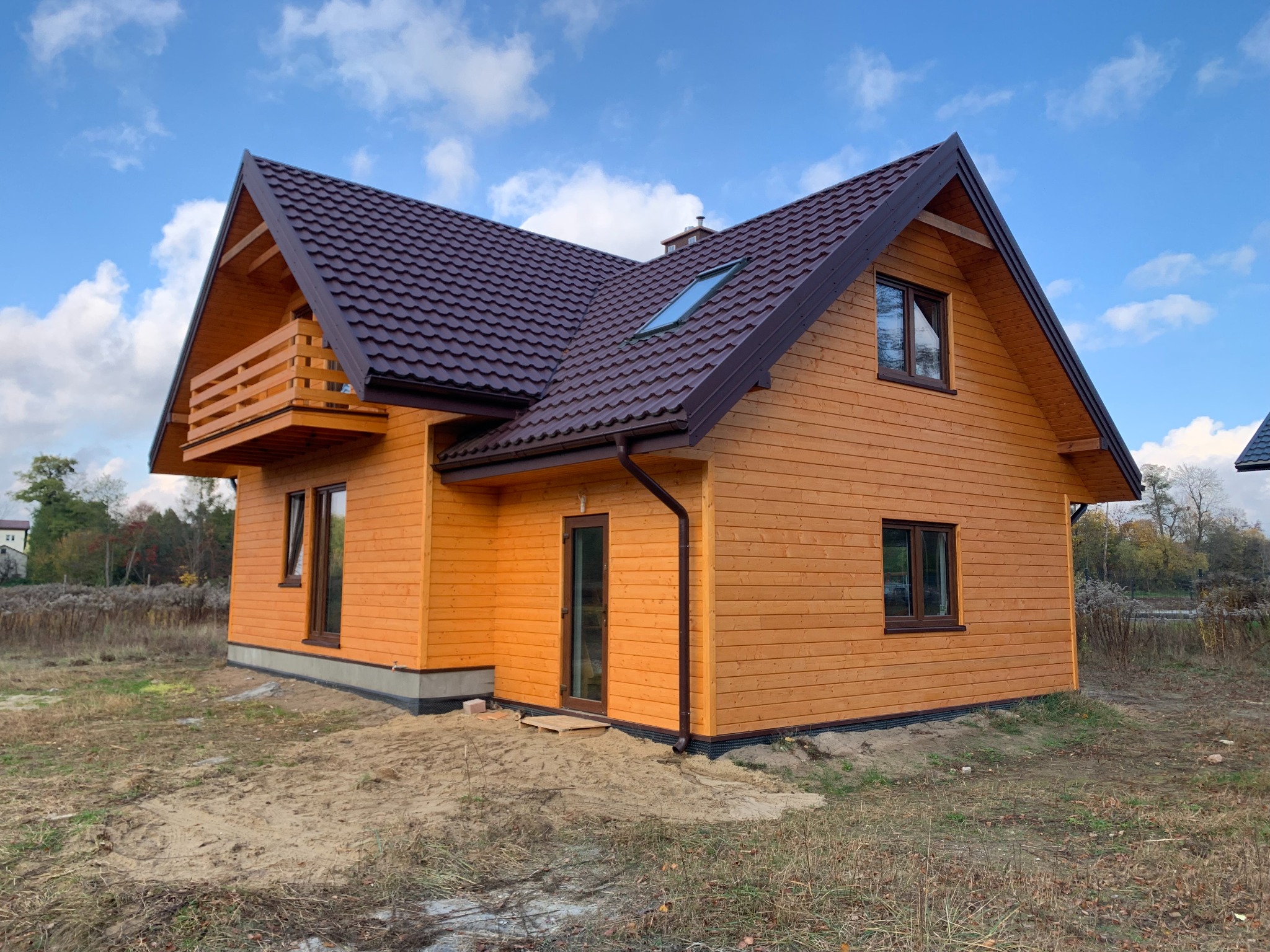 Nowoczesny, pomarańczowy dom z drewna z ciemnobrązowym dachem, balkonem i oknem dachowym, na działce z piaskiem i trawą.