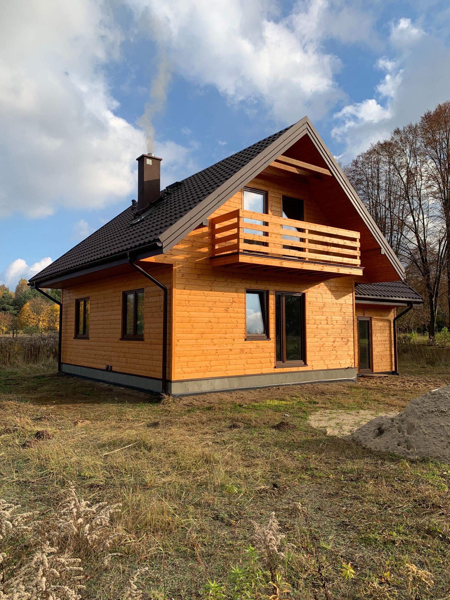 Nowoczesny dom szkieletowy z drewnianą elewacją i balkonem, z komina unosi się dym, otoczony jesiennym krajobrazem.