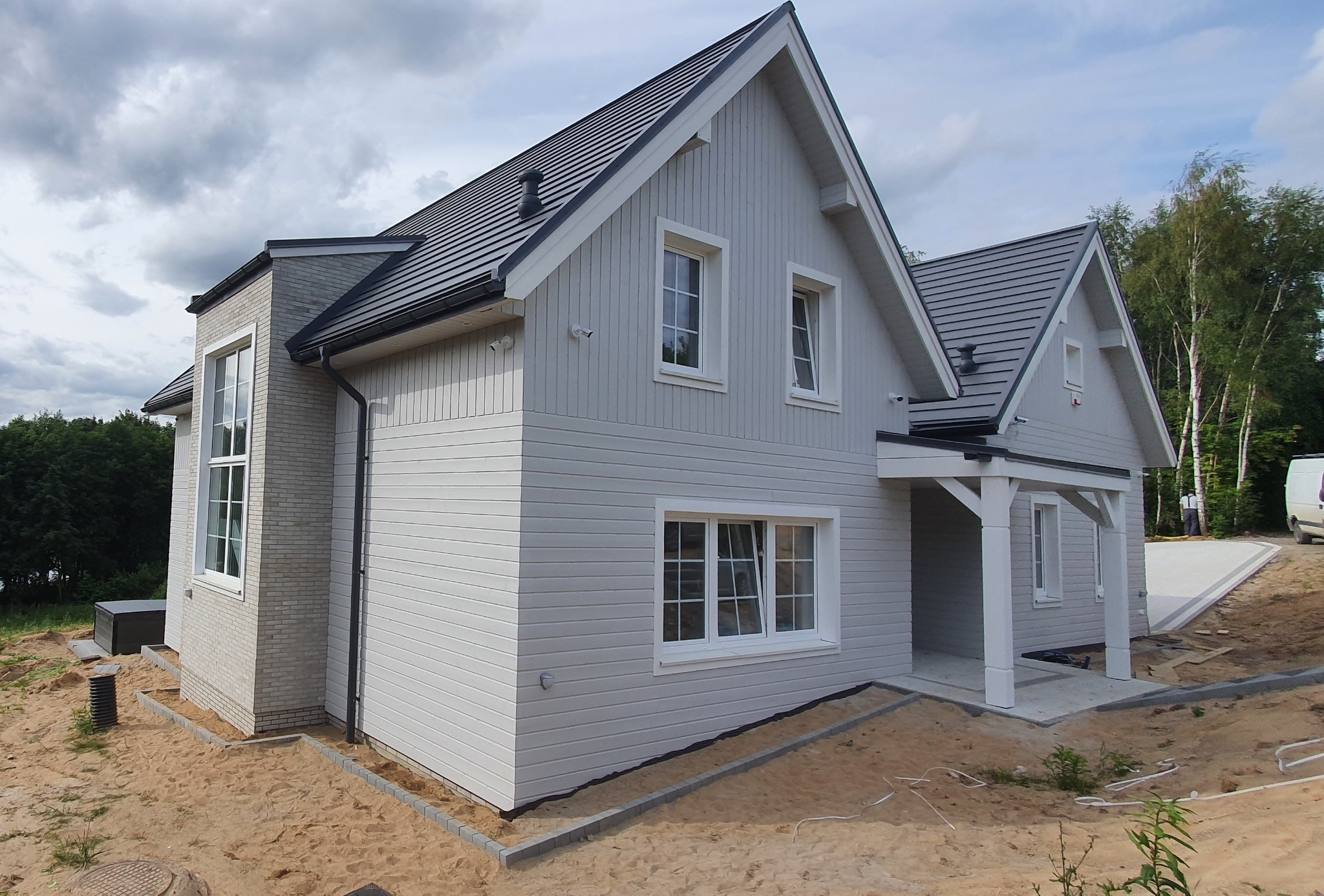 Jasnoszary dom z drewnianą elewacją i ciemnoszarą dachówką, z widocznym fragmentem dobudówki z cegły, w otoczeniu piaszczystego terenu i drzew.