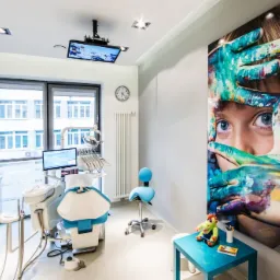 Jasne wnętrze gabinetu stomatologicznego z nowoczesnym fotelem dentystycznym, sprzętem medycznym, dużym oknem, zegarem na ścianie i kolorową grafiką z dzieckiem malującym rękami na ścianie...