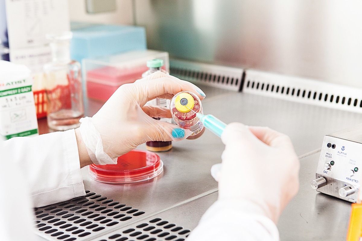 Pracownik w białym fartuchu i rękawiczkach laboratoryjnych pobiera próbkę z fiolki za pomocą strzykawki w sterylnym środowisku laboratoryjnym, obok widoczne płytki Petriego i aparatura pomiarowa.