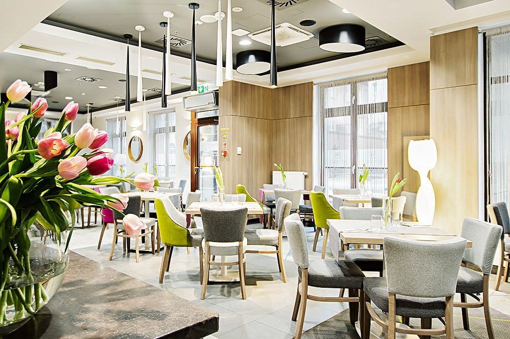 Wnętrze jasnej restauracji z bukietem tulipanów na pierwszym planie, minimalistyczne oświetlenie sufitowe i stoliki z krzesłami w odcieniach szarości i zieleni.