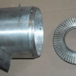 Metalowy cylinder z przymocowanym wałem i ostrzem obok metalowego pierścienia z nacięciami, widoczne ślady obróbki.