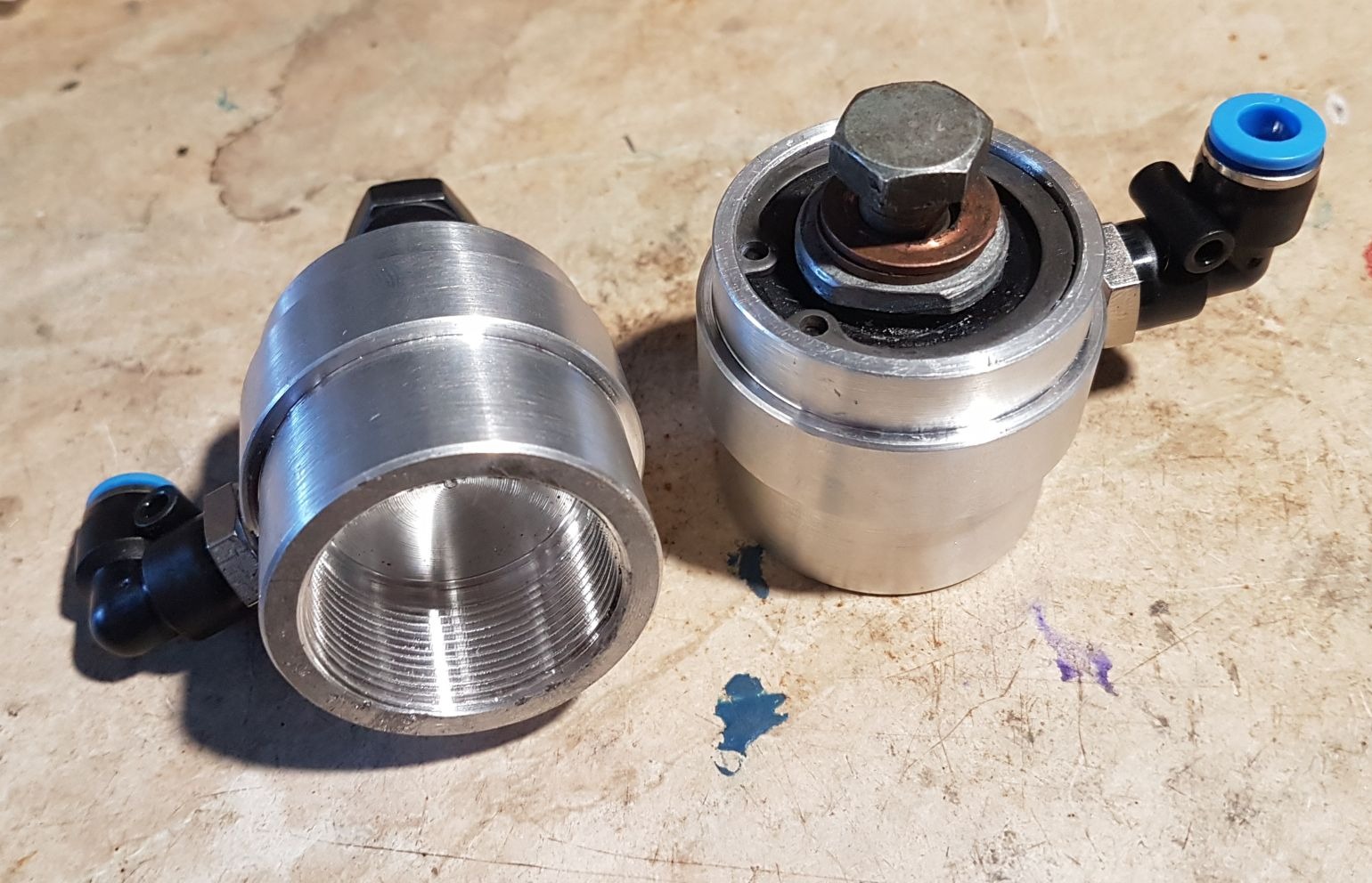 Dwa precyzyjnie obrobione, aluminiowe elementy z gwintami wewnętrznymi i złączkami pneumatycznymi, leżące na brudnym blacie roboczym. Jeden element z nakrętką i podkładkami na górze.