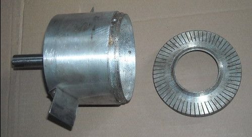 Metalowy cylinder z przymocowanym wałem i ostrzem obok metalowego pierścienia z nacięciami, widoczne ślady obróbki.