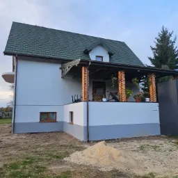 Dwukondygnacyjny dom z ocieploną elewacją, zielonym dachem z blachodachówki i werandą z ceglanymi kolumnami, widoczny stos piasku na trawniku.