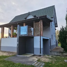 Dwukondygnacyjny dom z szarą elewacją, zielonym dachem i przeszkloną windą zewnętrzną na balkonie, widok z poziomu trawnika.