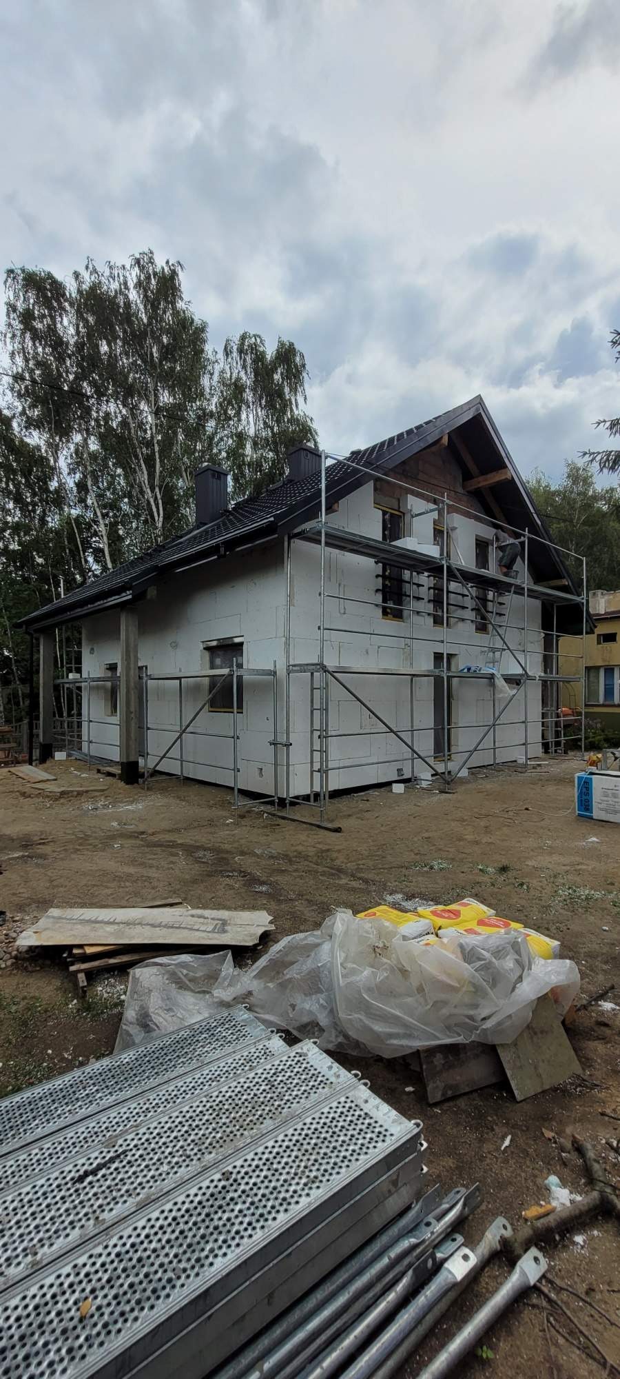 Dom jednorodzinny w trakcie ocieplania, widoczne rusztowania, styropianowe płyty elewacyjne i materiały budowlane na ziemi.