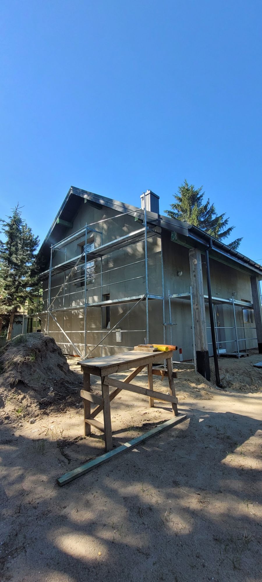 Dom jednorodzinny w trakcie prac elewacyjnych, otoczony rusztowaniem, z widocznym stołem roboczym i pryzmą ziemi na pierwszym planie.