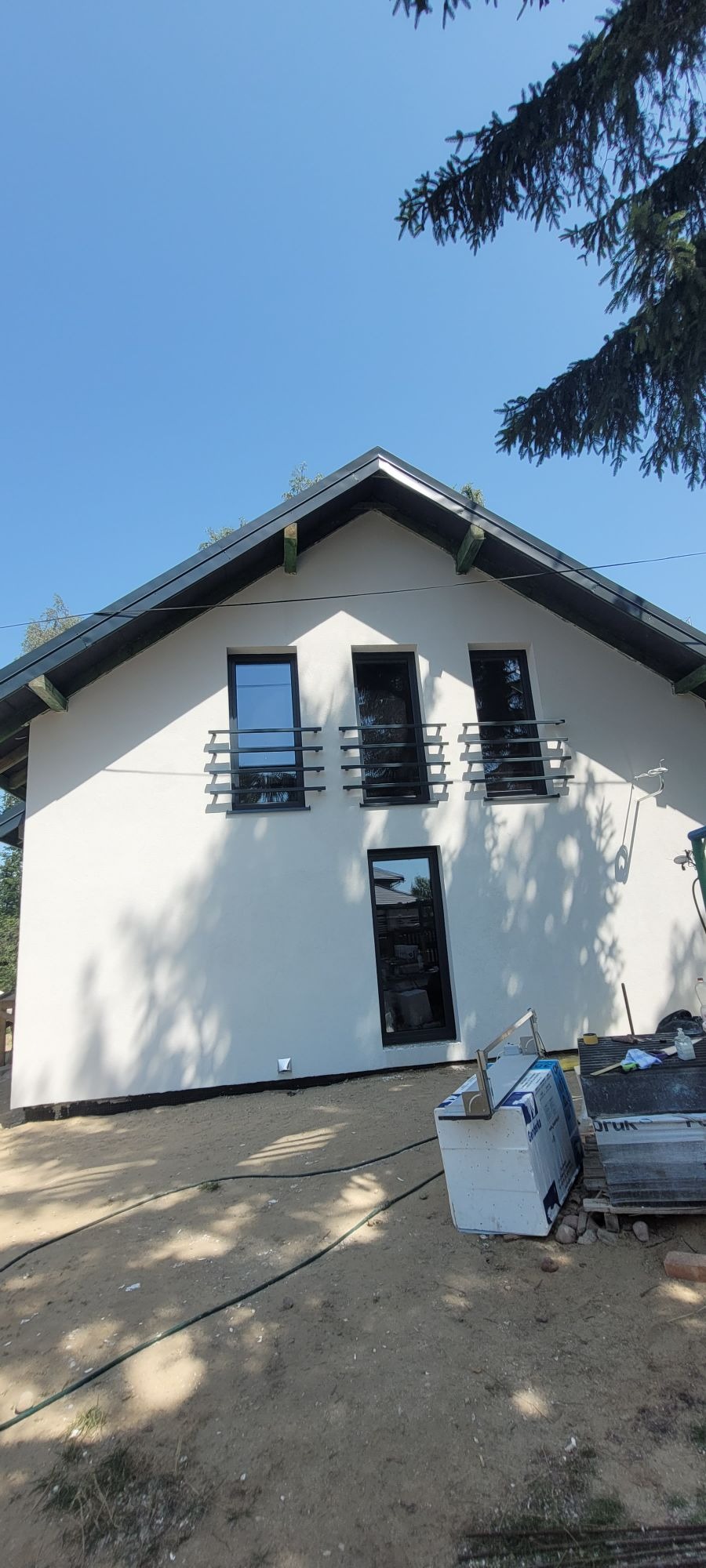 Świeżo otynkowany dom jednorodzinny z czarnymi oknami i ozdobnymi balustradami, widoczne materiały budowlane na posesji.