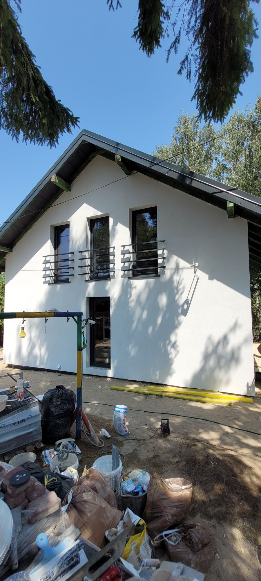 Biały dom jednorodzinny z czarnymi oknami i balustradami, po wykonaniu elewacji, widoczne materiały budowlane i narzędzia na pierwszym planie, drzewa w tle.