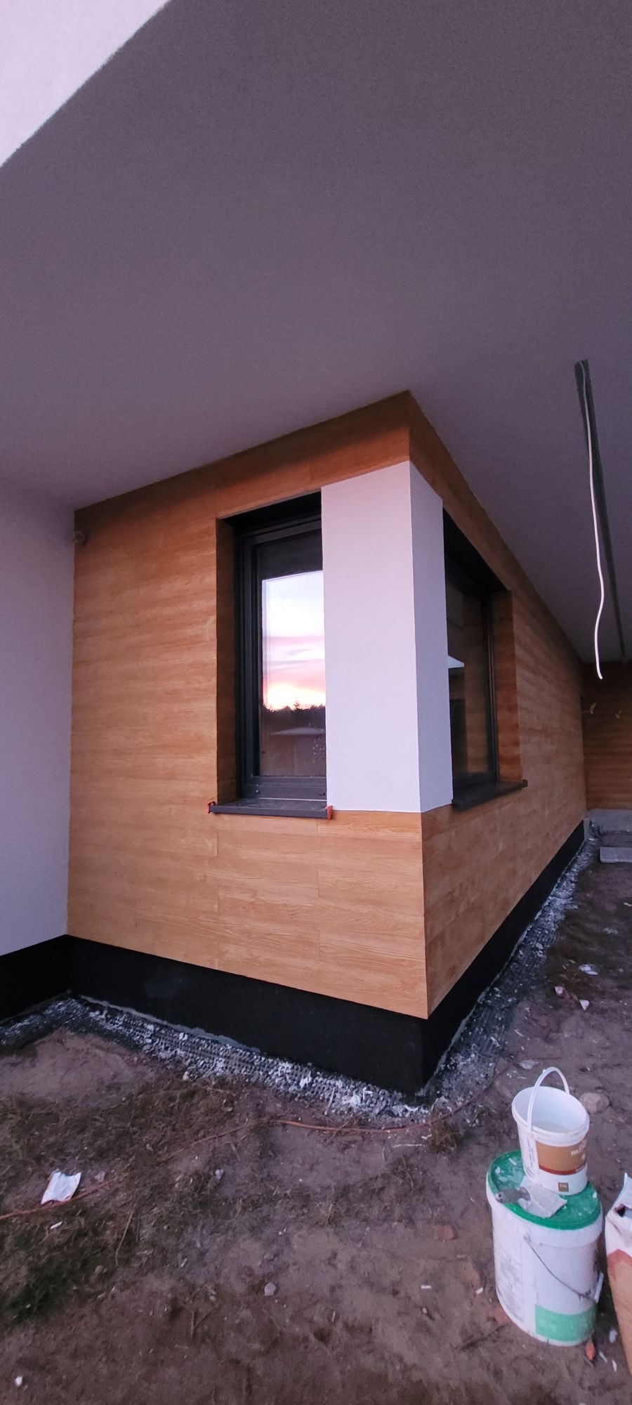 Fragment elewacji budynku z oknem, wykończonej panelami imitującymi drewno, widoczne detale wykończenia i dwa wiadra po farbie na ziemi.