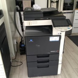 Copix Sp z o.o. - Używana kserokopiarka Konica Minolta bizhub C253 z trzema szufladami na papier, stojąca na drewnianej podłodze w pomieszczeniu biurowym, obok odkurzacz i inna kserokopiarka.