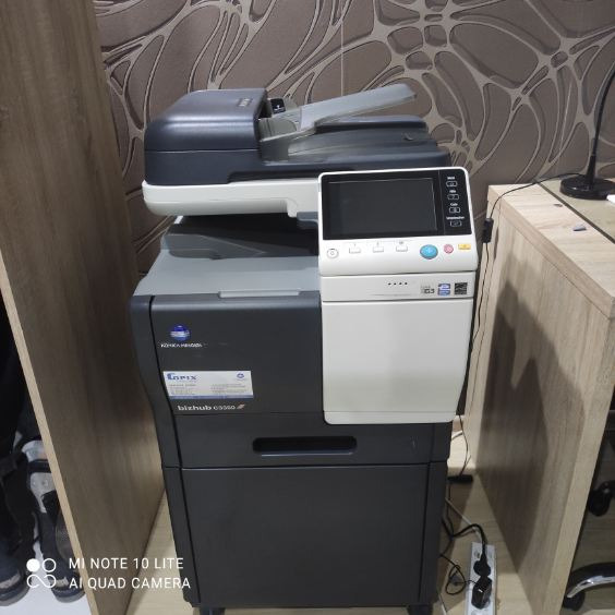 Używana kserokopiarka Konica Minolta bizhub C5550i, widoczna naklejka z modelem, ustawiona w biurze obok drewnianego biurka i ściany z wzorzystą tapetą.