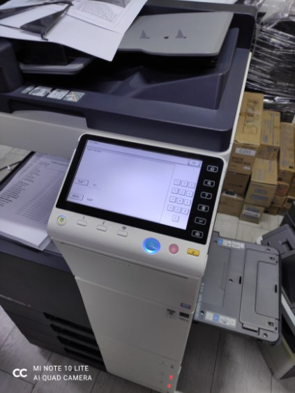 Używana kserokopiarka wielkoformatowa z otwartą szufladą na papier i stosem dokumentów na podajniku, widoczny ekran dotykowy i panel sterowania.