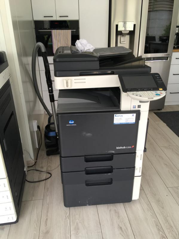 Używana kserokopiarka Konica Minolta bizhub C253 z trzema szufladami na papier, stojąca na drewnianej podłodze w pomieszczeniu biurowym, obok odkurzacz i inna kserokopiarka.