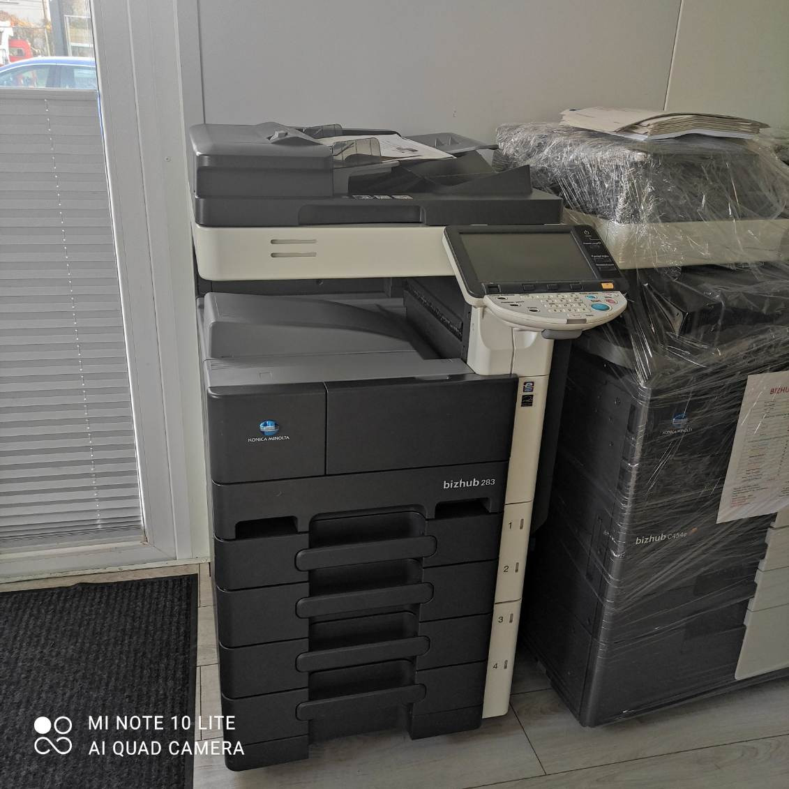 Używana kserokopiarka Konica Minolta bizhub 283 obok zapakowanej w folię kserokopiarki bizhub C454e, widok z boku, z numeracją szuflad na obudowie.