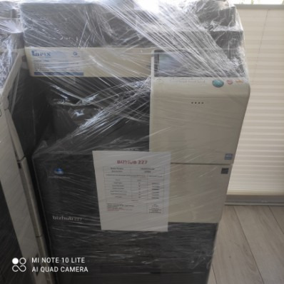 Używana kserokopiarka Konica Minolta bizhub 227, owinięta folią transportową, z widoczną specyfikacją techniczną przyklejoną z przodu.