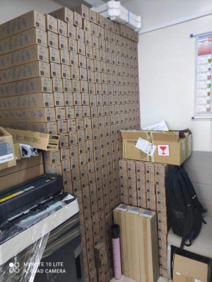 Stos kartonów z kserokopiarkami, magazyn z zapasem urządzeń biurowych, widoczna kserokopiarka owinięta folią, ściana kartonów sięgająca sufitu.