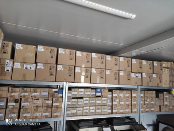 Rzędy kartonowych pudeł z etykietami na metalowych regałach w magazynie, widoczne modele i oznaczenia na pudełkach. Część pudeł zawiera sprzęt biurowy.