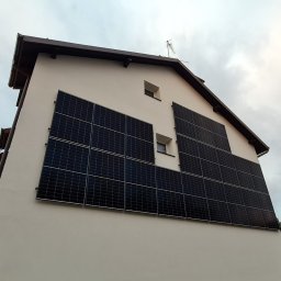 NNF ENERGY SPÓŁKA Z OGRANICZONĄ ODPOWIEDZIALNOŚCIĄ - Biały dom z panelami słonecznymi na ścianie bocznej, widoczne okna i antena na dachu, pochmurne niebo w tle. Nietypowy montaż paneli.