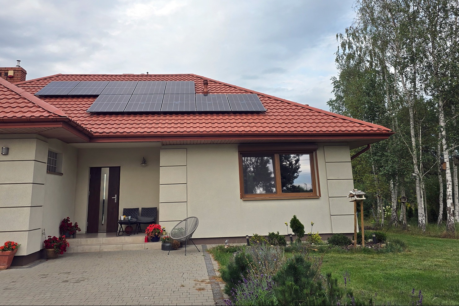 Dom jednorodzinny z panelami fotowoltaicznymi na czerwonym dachu. Widok z przodu, z ogrodem i drzewami w tle. Słoneczny dzień, błękitne niebo.