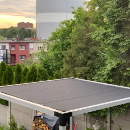 Nowoczesna pergola z płaskim, ciemnym dachem na tle zieleni, budynków mieszkalnych i zachodzącego słońca.