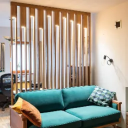 Turkusowa sofa z drewnianą ramą i poduszkami, na tle drewnianej ścianki działowej z wbudowanym oświetleniem LED.