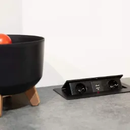 Czarny, minimalistyczny blat z wbudowanym gniazdem elektrycznym i portami USB, obok czarna misa na trzech nóżkach z owocami.