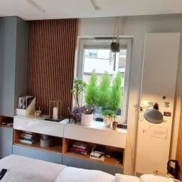 Sypialnia z zabudową meblową w odcieniach szarości i drewna, z lamelami na ścianie za łóżkiem i przy oknie, oraz roślinami w doniczkach na parapecie.